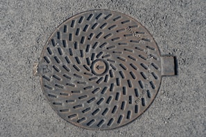 A round metal manhole cover is set in asphalt. The cover features a spiral pattern of slots and has text engraved on it. The surface shows signs of wear with a slightly weathered appearance.
