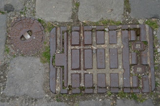 A rectangular metal manhole cover with raised rectangles and text, placed on a cobblestone pavement. Next to it is a round metal access lid with a small white handle. Small patches of grass are growing between the stones and manhole cover.