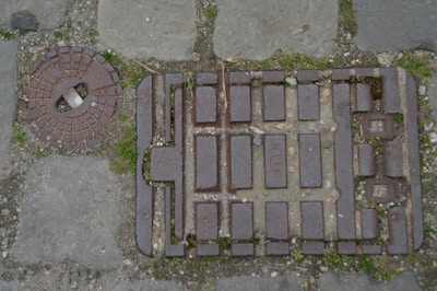 A rectangular metal manhole cover with raised rectangles and text, placed on a cobblestone pavement. Next to it is a round metal access lid with a small white handle. Small patches of grass are growing between the stones and manhole cover.