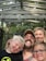 A family of four is smiling and posing for a selfie outdoors. They are standing under a wooden structure with the words 'LET NATURE BE YOUR TEACHER' written on it. The background is filled with green trees.