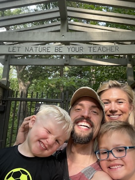 A family of four is smiling and posing for a selfie outdoors. They are standing under a wooden structure with the words 'LET NATURE BE YOUR TEACHER' written on it. The background is filled with green trees.