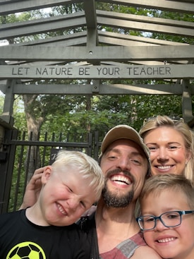 A family of four is smiling and posing for a selfie outdoors. They are standing under a wooden structure with the words 'LET NATURE BE YOUR TEACHER' written on it. The background is filled with green trees.