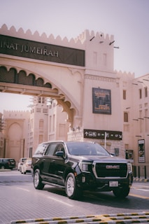 A black GMC SUV is parked in front of an architectural structure with Middle Eastern design elements, including arched windows and geometric patterns. Signage and decorative elements are visible, with a background indicating a modern shopping or cultural area.