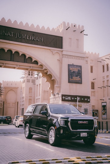 A black GMC SUV is parked in front of an architectural structure with Middle Eastern design elements, including arched windows and geometric patterns. Signage and decorative elements are visible, with a background indicating a modern shopping or cultural area.