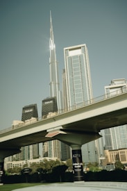 A modern cityscape features a prominent skyscraper, with other high-rise buildings surrounding it. In the foreground, an elevated section of a highway or metro line is visible, supported by sturdy columns. The scene conveys urban development and architectural innovation.