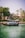 A small boat with a canopy floats on a calm, emerald green waterway, carrying several passengers who are seated. In the background, palm trees and lush vegetation border a building with traditional architecture, creating a serene and leisurely scene.
