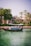 A small boat with a canopy floats on a calm, emerald green waterway, carrying several passengers who are seated. In the background, palm trees and lush vegetation border a building with traditional architecture, creating a serene and leisurely scene.