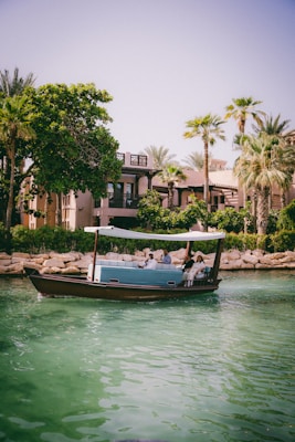A small boat with a canopy floats on a calm, emerald green waterway, carrying several passengers who are seated. In the background, palm trees and lush vegetation border a building with traditional architecture, creating a serene and leisurely scene.