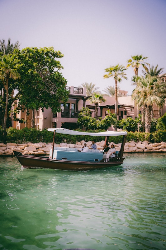 A small boat with a canopy floats on a calm, emerald green waterway, carrying several passengers who are seated. In the background, palm trees and lush vegetation border a building with traditional architecture, creating a serene and leisurely scene.