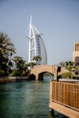 A modern, sail-shaped skyscraper stands prominently against a clear blue sky. In the foreground, a stone arch bridge spans a turquoise canal, bordered by lush palm trees and wooden railings. The scene conveys a blend of modern architecture and natural beauty.