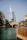 A modern, sail-shaped skyscraper stands prominently against a clear blue sky. In the foreground, a stone arch bridge spans a turquoise canal, bordered by lush palm trees and wooden railings. The scene conveys a blend of modern architecture and natural beauty.
