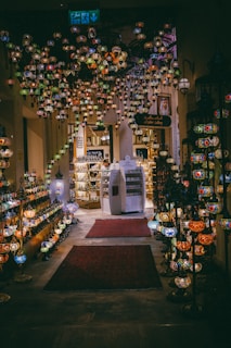 An interior view of a shop adorned with numerous colorful, hanging mosaic lanterns. The lanterns create a vibrant and ornate canopy, casting colorful reflections around the space. The shop features intricately designed carpets lining the floor, and shelves filled with an array of similar decorative items. The ambient lighting enhances the rich, warm tones of the decor.