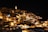 An ancient cityscape at night, illuminated by warm yellow lights that highlight the historic architecture and clustered buildings. The structures are made of stone and have a rustic quality, with a prominent tower lit up, standing out against the dark sky.