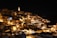 An ancient cityscape at night, illuminated by warm yellow lights that highlight the historic architecture and clustered buildings. The structures are made of stone and have a rustic quality, with a prominent tower lit up, standing out against the dark sky.