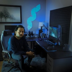 A person is seated on a gaming chair in a dimly lit room with a modern setup that includes a computer, microphone, and multiple screens. The walls are decorated with soundproofing panels and a geometric LED light panel. A framed picture of trees is hung on the wall. The atmosphere suggests a gaming or streaming environment.