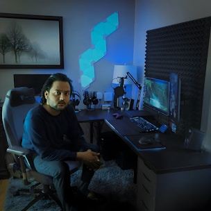 A person is seated on a gaming chair in a dimly lit room with a modern setup that includes a computer, microphone, and multiple screens. The walls are decorated with soundproofing panels and a geometric LED light panel. A framed picture of trees is hung on the wall. The atmosphere suggests a gaming or streaming environment.