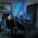 A person is seated on a gaming chair in a dimly lit room with a modern setup that includes a computer, microphone, and multiple screens. The walls are decorated with soundproofing panels and a geometric LED light panel. A framed picture of trees is hung on the wall. The atmosphere suggests a gaming or streaming environment.