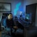 A person is seated on a gaming chair in a dimly lit room with a modern setup that includes a computer, microphone, and multiple screens. The walls are decorated with soundproofing panels and a geometric LED light panel. A framed picture of trees is hung on the wall. The atmosphere suggests a gaming or streaming environment.