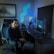 A person is seated on a gaming chair in a dimly lit room with a modern setup that includes a computer, microphone, and multiple screens. The walls are decorated with soundproofing panels and a geometric LED light panel. A framed picture of trees is hung on the wall. The atmosphere suggests a gaming or streaming environment.