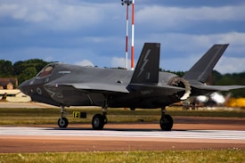 A sleek, modern fighter jet prepares for takeoff on a runway. It has a distinctive dark gray color with sharp angles and markings, suggesting advanced technology and military design. The background features a clear sky with clouds and a lush, green treeline.