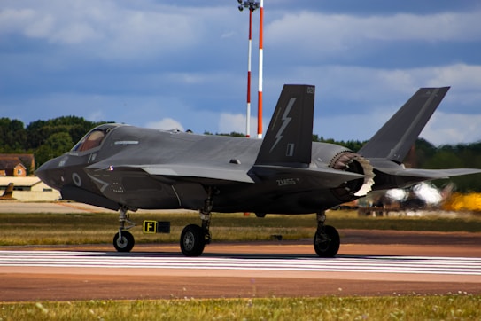 A sleek, modern fighter jet prepares for takeoff on a runway. It has a distinctive dark gray color with sharp angles and markings, suggesting advanced technology and military design. The background features a clear sky with clouds and a lush, green treeline.