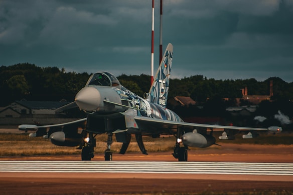 A military jet with a detailed camouflage paint design is on a runway. The jet's nose is pointed straight ahead, and the background includes a forested area and some industrial buildings under a cloudy sky.