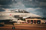 A modern fighter jet is positioned on a runway, with its pilot visible through the cockpit canopy. The airplane exhibits military markings and insignia, with its sleek design emphasized by the sunlight hitting the metallic surface. In the background, hangars and buildings are seen under a cloudy sky, contributing to an intense atmosphere.