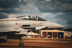 A modern fighter jet is positioned on a runway, with its pilot visible through the cockpit canopy. The airplane exhibits military markings and insignia, with its sleek design emphasized by the sunlight hitting the metallic surface. In the background, hangars and buildings are seen under a cloudy sky, contributing to an intense atmosphere.