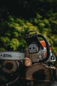 A chainsaw rests on a stack of cut logs. The chainsaw is prominently displayed with branding visible, surrounded by a blurred background of green foliage.