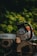 A chainsaw rests on a stack of cut logs. The chainsaw is prominently displayed with branding visible, surrounded by a blurred background of green foliage.