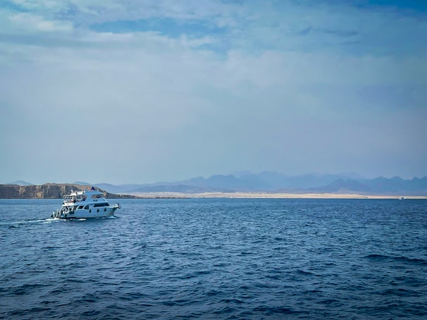 A small yacht navigates through calm waters with a distant desert landscape and mountains visible on the horizon under a partly cloudy sky.
