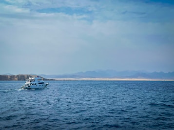 A small yacht navigates through calm waters with a distant desert landscape and mountains visible on the horizon under a partly cloudy sky.