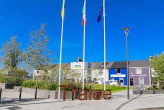 Three flagpoles stand prominently in front of a well-maintained urban setting with a sign that reads 'Tralee' in large letters. Behind the flagpoles, there are buildings with signs, one reading 'Foley & Associates' and the other 'community k'. Paved walking paths, green landscaping, and a parked SUV are visible, along with a bike rack.