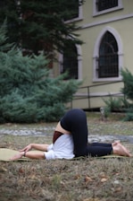 A person is practicing yoga outdoors, performing a pose on a mat placed on grass. They are wearing a white top and black leggings. The background features a building with arched windows and surrounding greenery.