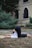 A person is practicing yoga outdoors, performing a pose on a mat placed on grass. They are wearing a white top and black leggings. The background features a building with arched windows and surrounding greenery.