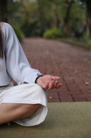 A person is sitting cross-legged on a green mat with their hand resting on their knee in a meditative pose. The setting is outdoors on a brick pathway lined with greenery, creating a peaceful atmosphere.