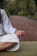 A person is sitting cross-legged on a green mat with their hand resting on their knee in a meditative pose. The setting is outdoors on a brick pathway lined with greenery, creating a peaceful atmosphere.