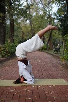 A person is balancing on their head in a yoga pose on a green yoga mat placed on a red brick walkway. The surrounding environment is a peaceful park with trees and green foliage, suggesting an outdoor setting. The person is dressed in loose-fitting white clothing, likely for comfort and flexibility.