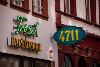 The image displays a building facade with two distinct signs. One sign features the text 'Frosch' in green neon script above 'PARFUMERIE' in yellow block letters. Another sign has '4711' in yellow against a teal background. The facade is primarily white with brown accents, and there are windows with red-brown trim visible above the signs.