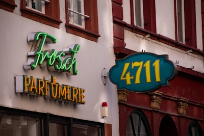 The image displays a building facade with two distinct signs. One sign features the text 'Frosch' in green neon script above 'PARFUMERIE' in yellow block letters. Another sign has '4711' in yellow against a teal background. The facade is primarily white with brown accents, and there are windows with red-brown trim visible above the signs.