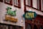 The image displays a building facade with two distinct signs. One sign features the text 'Frosch' in green neon script above 'PARFUMERIE' in yellow block letters. Another sign has '4711' in yellow against a teal background. The facade is primarily white with brown accents, and there are windows with red-brown trim visible above the signs.