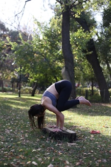 A person is practicing yoga outdoors, performing an advanced balancing pose on a tree stump in a park. The setting is lush with green grass and tall trees, and fallen leaves are scattered across the ground. The person is wearing athletic gear, consisting of a tank top and leggings.