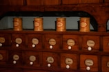 A vintage wooden apothecary cabinet with multiple drawers, each labeled with the name of a different substance or herb. On top of the cabinet are several cylindrical containers with labels, possibly used for storing various medicinal ingredients.
