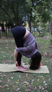 A person is performing a forward bend yoga pose on a mat in an outdoor setting surrounded by trees and grass. They are wearing a striped shirt and black leggings.