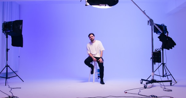 A man sits on a stool in a professional photography studio with soft, ambient blue lighting. He appears relaxed and is surrounded by various lighting equipment on stands. The background is plain and softly illuminated, creating a calm, serene atmosphere.