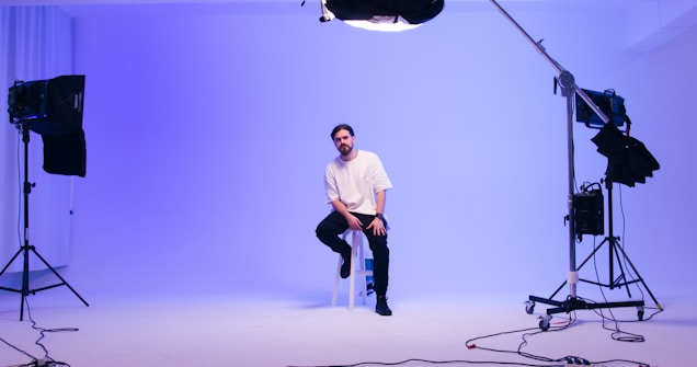 A man sits on a stool in a professional photography studio with soft, ambient blue lighting. He appears relaxed and is surrounded by various lighting equipment on stands. The background is plain and softly illuminated, creating a calm, serene atmosphere.