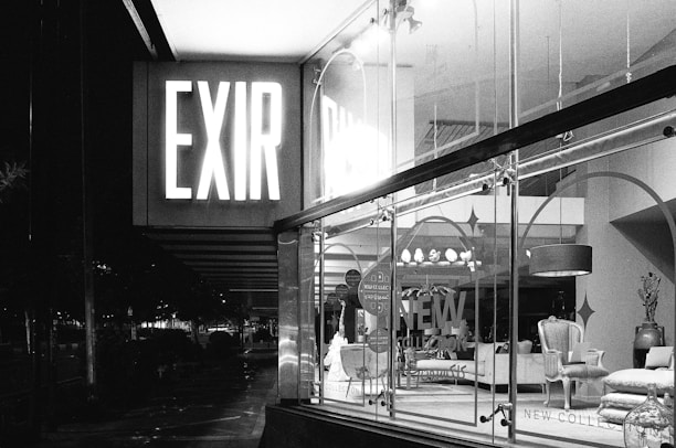 A black and white photograph depicts a store display with large glass windows. Illuminated signage reads 'EXIR' and the interior showcases elegant furniture, including chairs and couches. The setting appears to be a night scene.