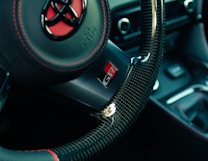 A close-up view of a car's steering wheel made of carbon fiber material with red stitching detailing. The steering wheel features a red and black logo in the center, with the letters GR on the silver part just below the center. The interior car elements are visible in the background, including parts of the dashboard and air vents.