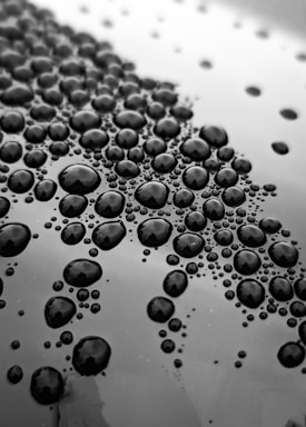 Multiple water droplets of varying sizes are scattered across a smooth, reflective surface, creating a pattern. The droplets are arranged closely together, displaying a high level of detail and clarity.
