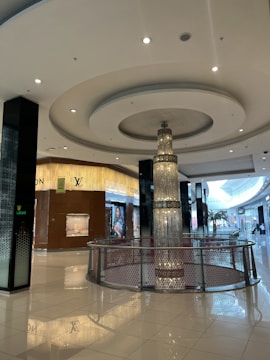 A luxurious indoor shopping mall featuring high-end stores such as Louis Vuitton. The mall has glossy tiled floors, a large central chandelier descending from the ceiling, and plenty of ambient lighting. The decor is modern and sophisticated, with clean lines and a spacious layout.